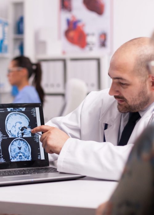 Brain surgeon pointing at ct scan on laptop in hospital office discussing with elderly patient about intervention. Nurse holding x-ray image in the background. Young sick woman in clinic corridor.